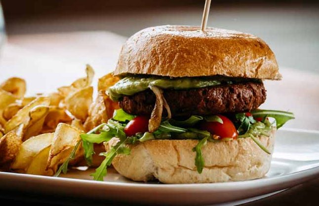 Veganer Burger mit Salat, Tomaten und einer grünen Avocado-Sauce, serviert mit Kartoffelspalten.