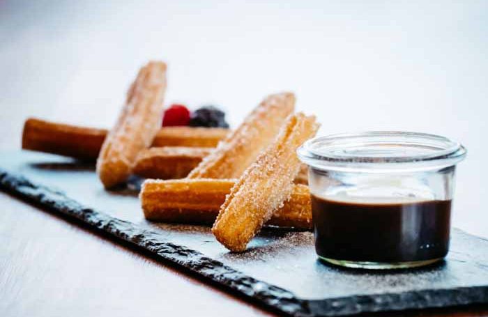 Churros mit Schokoladensauce und Beeren auf einem schwarzen Teller.