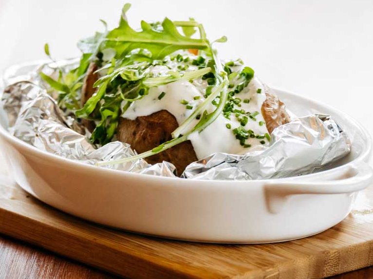 Ein gebackener Kartoffel mit Sauerrahm, Kräutern und frischem Rucola auf einem Holzbrett.